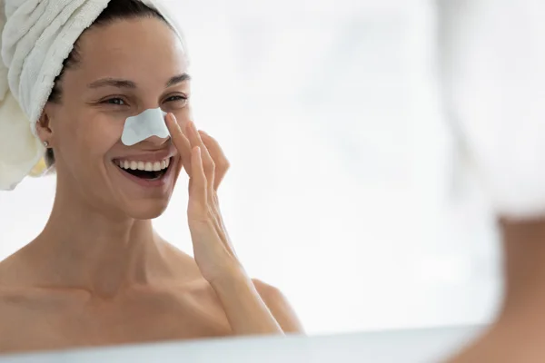 Młoda roześmiana kobieta w plastrem na nosie przegląda się w lustrze